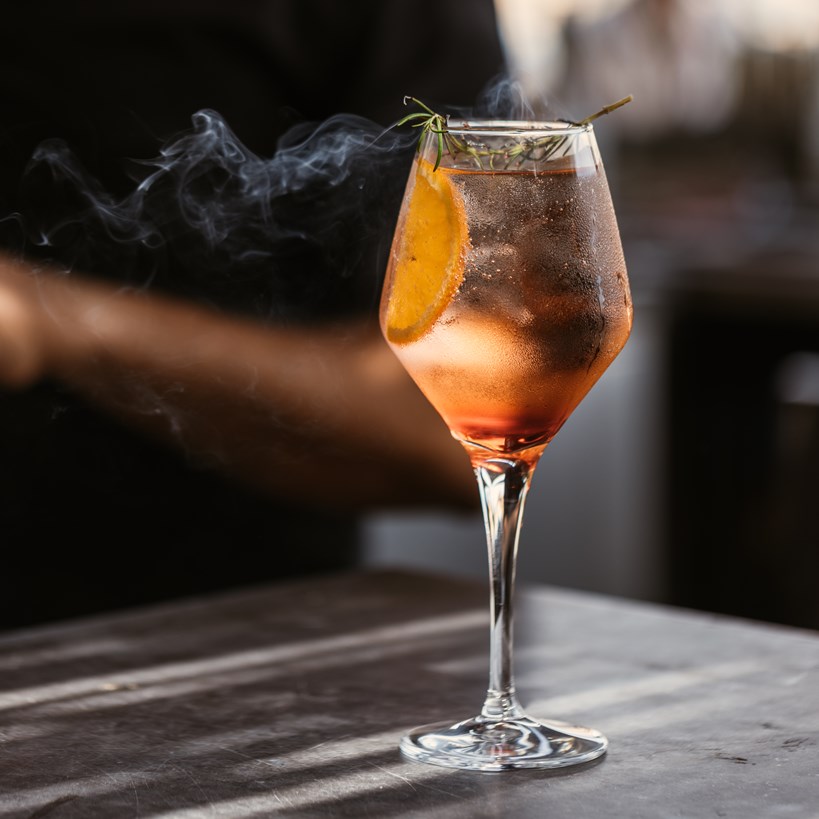 Cocktail in a wine glass with a slice of orange and essence of burnt rosemary